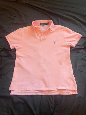 Rosa pikétröja från Polo Ralph Lauren - Säljer en rosa pikétröja från Polo Ralph Lauren i slim fit-modell. Tröjan har korta ärmar, klassisk krage och knappar vid halsen. Den ikoniska blå loggan är broderad på bröstet. Perfekt för en avslappnad men snygg stil.