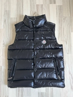 Svart dunväst från Moncler - Snygg svart dunväst från Moncler med glansig finish, hög krage och två dragkedjefickor framtill. Västen har Moncler-logga på bröstet och stängs med knappar. Perfekt för lager-på-lager och streetstyle.