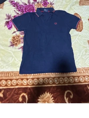 Marinblå pikétröja från Fred Perry - Klassisk marinblå pikétröja från Fred Perry med röd och vit detalj på krage och ärmslut. Tröjan har korta ärmar, knappslå och den ikoniska Fred Perry-loggan broderad på bröstet. Perfekt till jeans eller kjol för en sportig look.