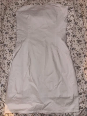 Vit tube kort klänning - Stilren vit axelbandslös kort klänning med figurnära passform. Klänningen har en slit på baksidan. 