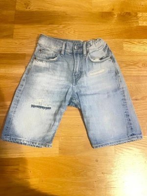 Ljusblå denimshorts med slitningar - Snygga ljusblå jeansshorts i denim med slitna detaljer och lagningar framtill. Klassisk femficksmodell med knapp och dragkedja. Perfekta för en avslappnad streetstil.