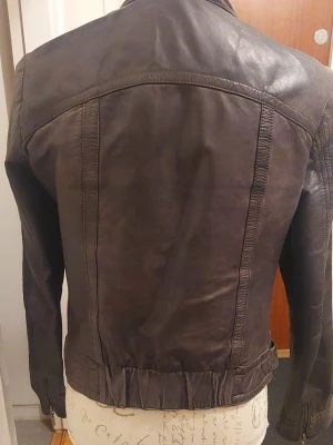 Mörkbrun skinnjacka med dragkedjor - Snygg svart, dock har färgen nöts och har lite bruna genomslag, skinnjacka i klassisk bikerstil med ståkrage och två dragkedjeförsedda bröstfickor. Jackan har figurnära passform och detaljerade sömmar längs ryggen och sidorna. 