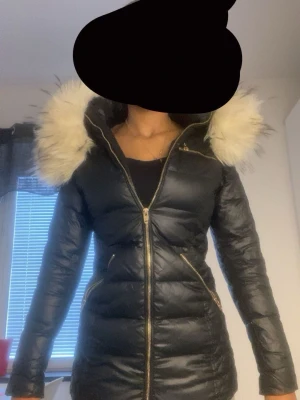 Svart lång dunjacka med fuskpäls - Snygg svart rocknblue  l från Frost med guldfärgade dragkedjor och detaljer. Jackan har en stor huva med fluffig fuskpäls och figurnära passform. Märkeslogga på ärmen och två snedställda fickor framtill. Perfekt för kalla vinterdagar fler bilder e ba o kontakta 💕