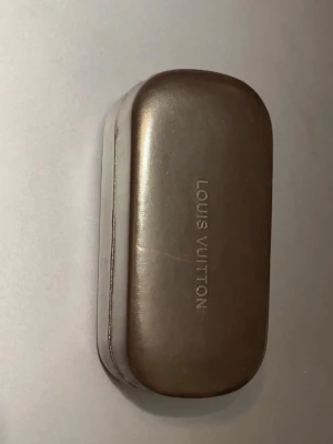 Lv glasögon - Snyggt solglasögonfodral från Lv i mörkbrunt skinn med ljus insida. Fodralet har en stilren, avlång form och loggan är tryckt både på utsidan och insidan. Perfekt för att skydda dina solglasögon med extra lyxig känsla.