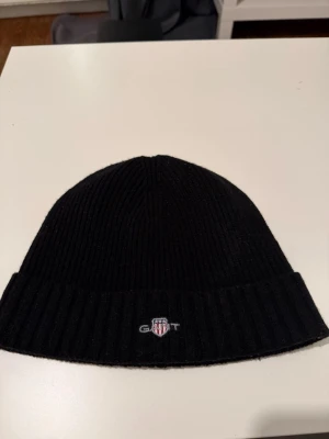 Svart ribbstickad mössa från GANT - Snygg svart ribbstickad mössa från GANT med broderad logga framtill. Klassisk modell med uppvikt kant och mjukt material som håller dig varm. Perfekt accessoar för kyliga dagar och enkel att matcha med olika outfits.