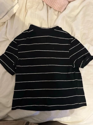 Svart t-shirt med vita ränder - En svart t-shirt med tunna vita horisontella ränder. T-shirten har korta ärmar och en klassisk rund halsringning. Passformen är något boxig och materialet ser ut att vara mjukt och bekvämt, perfekt för en avslappnad stil.