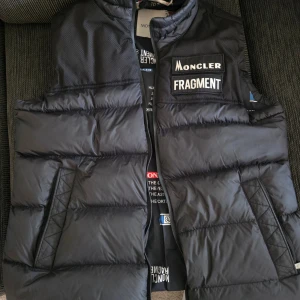 Svart dunväst Moncler Fragment - Säljer en svart dunväst från Moncler Fragment Västen har quiltad design, två snedställda fickor och tryckt innerfoder. 