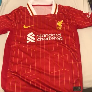 Liverpool FC röd matchtröja Nike - Liverpool FCs officiella matchtröja i rött med gula detaljer och tunna gula ränder. Broderad LFC-logga på bröstet, Nike swoosh och sponsortryck. Klassisk krage med vita och gula inslag. Tillverkad i lätt och ventilerande polyester.