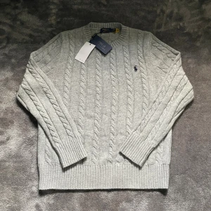 Grå kabelstickad tröja Polo Ralph Lauren - Kabelstickad grå tröja från Polo Ralph Lauren med klassisk rund halsringning och broderad logga på bröstet. Tröjan är långärmad och har breda ribbade muddar vid ärmslut och nederkant. Perfekt för lager-på-lager under kyliga dagar.