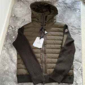 Olivgrön pufferjacka från Moncler - Säljer en olivgrön pufferjacka från Moncler med stickade mörkbruna ärmar och huva. Jackan har dragkedja framtill, Moncler-logga på ärmen och quiltad kropp i syntetmaterial. Perfekt för dig som vill ha en snygg och bekväm jacka med coola detaljer. Storlek L men passar M