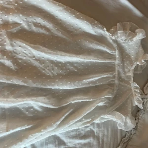 Vit blus med volangdetaljer - Supersöt vit blus med små prickar i tyget och volangdetaljer vid ärmslut och halsringning. Klänningen är ärmlös och har en luftig känsla, perfekt för varma dagar. Materialet känns lätt och andas, vilket gör den extra bekväm.