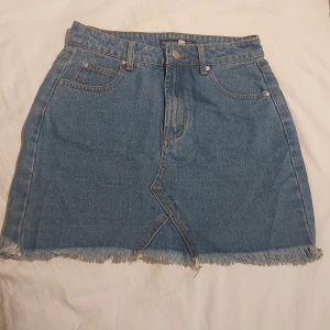 Blå jeanskjol från Boohoo, strl 34 - Snygg blå jeanskjol från Boohoo med fransad nederkant och klassiska fickor fram och bak. Kjolen har en cool omlott-detalj framtill.