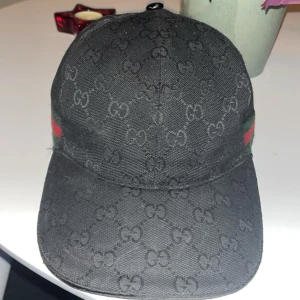 Svart Gucci keps med GG-mönster - Snygg svart keps från Gucci med klassiskt GG-monogram över hela kepsen och ikoniska gröna och röda ränder på sidan. Kepsen är tillverkad i bomull och har justerbar rem baktill för perfekt passform. Perfekt accessoar för en trendig streetstyle-look.