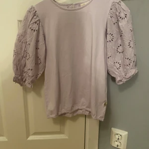 Ljuslila blus med puffärmar från Pompdelux - Supersöt ljuslila blus från Pompdelux med broderade puffärmar i bomull och rund halsringning. Ärmarna har fina broderade blommor och resår vid ärmslutet för extra volym. Perfekt för dig som gillar romantiska detaljer och en trendig look.