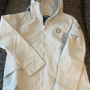 Ljusgrå vindjacka från Svea -Nyskick  - Snygg ljusgrå vindjacka från Svea med dragkedja, huva med snörning och två stora fickor framtill. Jackan har Svea-logga på bröstet och en broderad amerikansk flagga på ärmen. Perfekt för blåsiga dagar och enkel att matcha med allt.