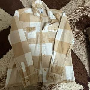 Beige och vit rutig overshirt - Snygg overshirt i beige och vitt rutigt mönster. Skjortan har två bröstfickor med knappar, klassisk krage och långa ärmar med manschett. Perfekt för lager-på-lager och en chill stil.