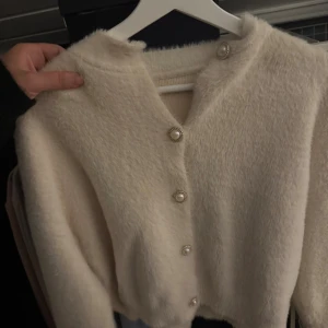 Beige fluffig kofta med pärlknappar - Supermjuk och fluffig beige kofta med snygga pärlknappar framtill. Koftan har lång ärm och en croppad passform som ger en trendig look. Perfekt att slänga på sig när det är kyligt ute.