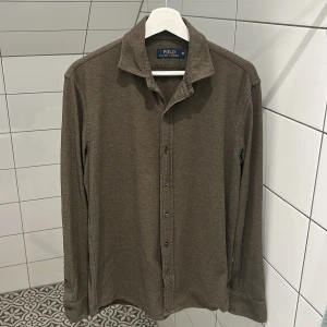  Polo Ralph Lauren - Säljer en brun långärmad skjorta från Polo Ralph Lauren. Skjortan har klassisk krage, knäppning framtill och är tillverkad i ett mjukt bomullstyg. Perfekt för en clean och avslappnad look. Storlek M