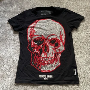 Svart t-shirt med strass från Philipp Plein - Svart t-shirt från Philipp Plein med glittrande strassdetaljer och ett stort tryck framtill i rött och silver. Klassisk passform med korta ärmar och rund hals. Liten metallbadge med logga på ryggen. Perfekt för dig som gillar statement-plagg.