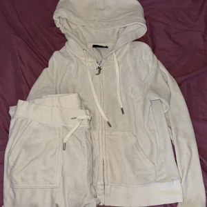 Beige Juicy Couture hoodie i velour - Supermjuk beige hoodie från Juicy Couture i klassisk velour med dragkedja och huva. Tröjan har snörning i halsen, långa ärmar och en snygg JC-logga broderad på bröstet. Perfekt för en chill och trendig look. ( byxorna är i storlek M o koftan S )