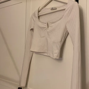 Vit croppad långärmad topp Sister - Säljer en vit croppad långärmad topp från Sister i storlek XXS. Toppen har ribbat tyg, diskret randigt mönster och en snygg v-ringning med liten slits framtill. Perfekt att styla med höga jeans eller kjol.