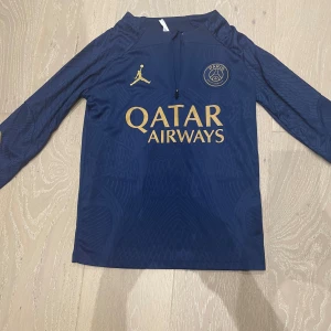 PSG x Jordan långärmad träningströja - Säljer en mörkblå långärmad PSG träningströja från Jordan med guldiga detaljer och loggor. Tröjan har dragkedja vid halsen och är tillverkad i ett lätt, sportigt material som andas. Matchande byxor medföljer, även de med PSG och Jordan-logga i guld. För barn runt 130-140