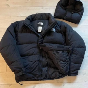 Svart dunjacka från The North Face - Säljer en svart dunjacka från The North Face med avtagbar huva och dragkedja. Jackan är quiltad och har justerbar dragsko nertill. Perfekt för kalla dagar och har en klassisk, oversized passform med lång ärm och diskret logga på bröstet.