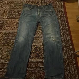 Levi's 501 blå jeans W32 L32 - Klassiska Levi's 501 jeans i blå tvätt med raka ben och knäppning fram. Jeansen har slitningar och hål på ena benet för en cool, avslappnad look. Tillverkade i bomull och passar perfekt för dig som gillar tidlös denimstil. 