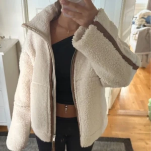Beige teddyjacka med bruna detaljer - Supermysig beige teddyjacka med bruna detaljer längs dragkedjan och ärmarna. Jackan har hög krage, två fickor framtill och stängs med dragkedja. Perfekt för kyliga dagar och ger en riktigt trendig look.