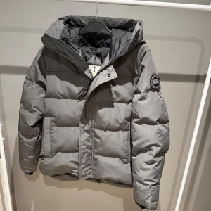Grå pufferjacka från Canada Goose - Säljer en grå pufferjacka från Canada Goose med huva och svart logotyp på ärmen. Jackan är vadderad och har dragkedja framtill samt stora fickor. Perfekt för kalla dagar och har en clean, modern look.