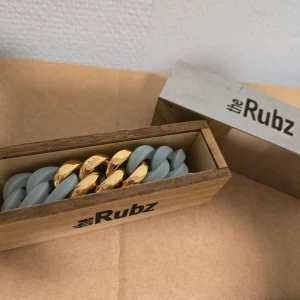 Blått och guldigt armband från The Rubz - Snyggt chunky armband från The Rubz med breda länkar i matt grått och glänsande guld. Kommer i original trälåda med logga. Perfekt statement-piece för dig som gillar att sticka ut med accessoarer.