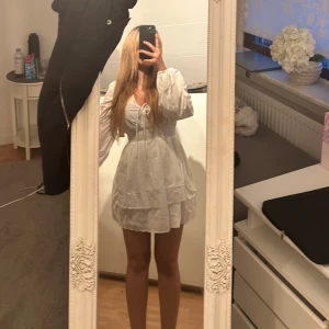 Vit kort klänning med volang - Supersöt vit kort klänning från Nelly med långa puffärmar, knytning framtill. Fin till sommarn och en perfekt studentklänning. Hade precis köpt den men är för tight. Provad som ni ser endast för bilden. Prislappen sitter kvar. Lär strykas