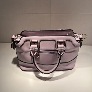 Ljusrosa handväska i skinnlook - Snygg ljusrosa handväska med svarta detaljer och guldfärgade metalldelar. Väskan har dubbla handtag, justerbar axelrem och dragkedja upptill. Insidan är fodrad i mörkbrunt tyg och har flera fack för småsaker. Perfekt för dig som gillar stilrena accessoarer.