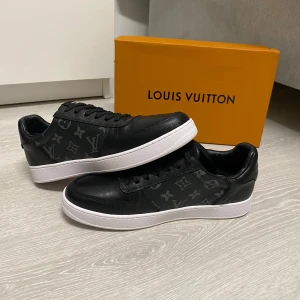 Louis Vuitton skor - Svarta Louis Vuitton sneakers i skinn med klassiskt monogrammönster i grått på sidorna. Skorna har vit platt sula, rund tå och snörning. Perfekta för dig som vill ha en lyxig och stilren look med streetkänsla.