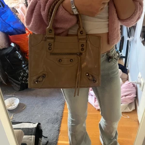 Beige handväska i skinn med dragkedja - Snygg beige handväska i skinn med coola detaljer och dragkedja framtill. Väskan har två rejäla handtag och ett rymligt huvudfack med innerficka. Perfekt storlek för att få plats med allt du behöver och passar till många olika outfits.