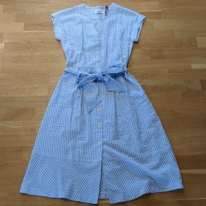 Blåvit rutig klänning Holly & Whyte XS - Supersöt blåvit rutig klänning från Holly & Whyte i 100% bomull. Klänningen har knappar framtill, v-ringning, korta ärmar och ett knytband i midjan. Perfekt för varma dagar med sin luftiga känsla och klassiska look.