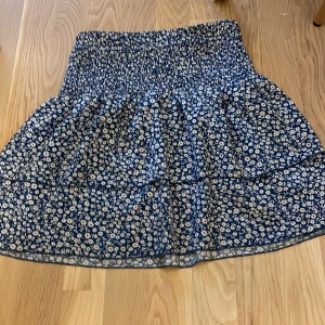 Blå blommig kortkjol från Emery Rose - Supersöt blå kortkjol från Emery Rose med vitt blommönster och smockad midja. Kjolen har en luftig och lätt form som ger en skön känsla. Perfekt för dig som gillar en feminin och trendig stil.