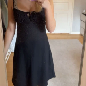 Svart kort klänning från Gina Tricot - Supersöt svart klänning från Gina Tricot med smala axelband och knytning framtill vid bysten. Klänningen har en enkel, A-linjeformad siluett och är perfekt för dig som gillar en stilren look. Materialet känns mjukt och lätt, perfekt för varmare dagar.
