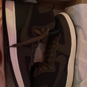 Nike Air Force 1 High svart/brun - Säljer ett par Nike Air Force 1 High i svart och mörkbrun med klassisk vit sula. Skorna har snörning och är höga i modellen, vilket ger en riktigt clean och stilren look. Ikonisk Swoosh på sidan och extra skosnören samt guld-detaljer medföljer.Dom är helt oanvända