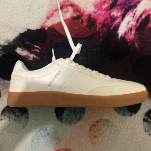 Vita sneakers från Jack & Jones - Snygga vita sneakers från Jack & Jones med klassisk design och bruna gummisulor. Skorna har snörning och är tillverkade i syntetmaterial med stilrena detaljer. Perfekta för dig som gillar en clean och sportig look.