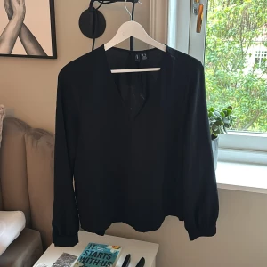 Svart blus från Vero Moda i M - Säljer en stilren svart blus från Vero Moda i storlek M. Blusen har v-ringning, långa ärmar med lätt puff vid ärmslut och är tillverkad i ett tunt, mjukt tyg. Perfekt att styla med jeans eller kjol för en clean look.