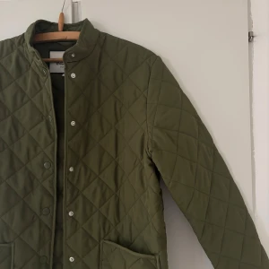 Olivgrön quiltad jacka från Indiska XS - Snygg olivgrön quiltad jacka från Indiska i storlek XS. Jackan har raka linjer, tryckknappar framtill, ståkrage och stora fickor. Perfekt för dig som gillar stilren och trendig look. Materialet är mjukt och lätt, passar bra till lager-på-lager. Inga tecken på användning!
