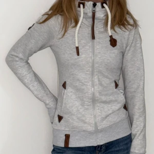 Ljusgrå hoodie från Naketano  - Ljusgrå hoodie från Naketano med dragkedja, stora vita snören i huvan och bruna detaljer i läderimitation. Snyggt emblem med uggla på ärmen och ribbade muddar.            Super mjuk utan noppor.