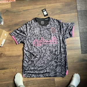 FC Palestina svart/grå fotbollströja XL - Unik fotbollströja från FC Palestina i svart och grått med blommigt mönster och rosa detaljer. Tryckt FC Palestina-märke på bröstet och rosa arabiskt tryck över bröstet. Tillverkad i lätt och ventilerande material, perfekt för match eller träning.