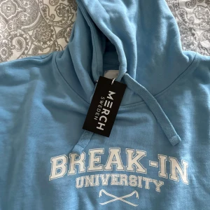 Ljusblå hoodie från Merch Sweden  - Babyblå hoodie från Merch Sweden med trycket 'Break-In University' på bröstet. Tröjan har huva med snörning, magficka och är tillverkad i 100% bomull. Helt ny med prislapp, aldrig andvänd. Stolek xl men passar L och m ifall man vill ha den overzize!