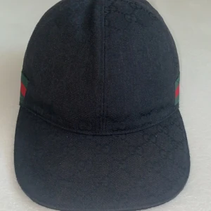 Gucci keps svart - Svart keps från Gucci i bomull med diskret GG-monogram över hela ytan. Klassisk form med böjd skärm och justerbar rem baktill. Sidorna har de ikoniska gröna och röda ränderna. Perfekt för dig som vill ha en stilren och lyxig accessoar.