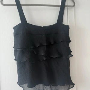 Svart volangtop från By Malene Birger - Supersnygg svart topp från By Malene Birger med breda axelband och flera lager volanger. Toppen har knappar bak och är gjord i ett lätt, plisserat tyg som ger en cool struktur. Sömmen har gått upp där bak på bilden som syns men går lätt att sy!