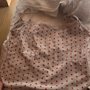 Vit linnetopp med röda hjärtan - Supersöt vit linnetopp med tunna axelband och röda hjärtan över hela tyget. Toppen har en liten spetskant upptill och en söt rosett i mitten. Perfekt för dig som gillar gulliga detaljer och romantisk stil.