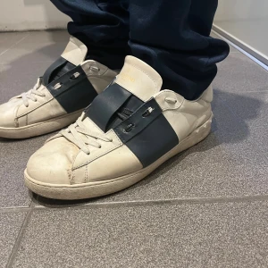 Valentino Garavani Open vit/marinblå - Snygga Valentino Garavani Open sneakers i vitt skinn med bred marinblå läderdetalj över mitten. Klassisk rund tå, platt sula och coola nitar baktill på hälen. Skorna har snörning och en stilren design som sticker ut. Perfekta för dig som gillar exklusiva streetwear-vibbar.