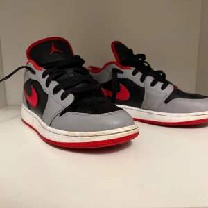 Nike Air Jordan 1 Low svart/röd/grå - Snygga Nike Air Jordan 1 Low sneakers i svart, grått och rött. Skorna har klassisk röd swoosh, svart tå och gråa paneler. Röd yttersula och detaljer på plösen. Tillverkade i skinn med svarta skosnören och låg profil. Perfekta för dig som gillar streetstyle.